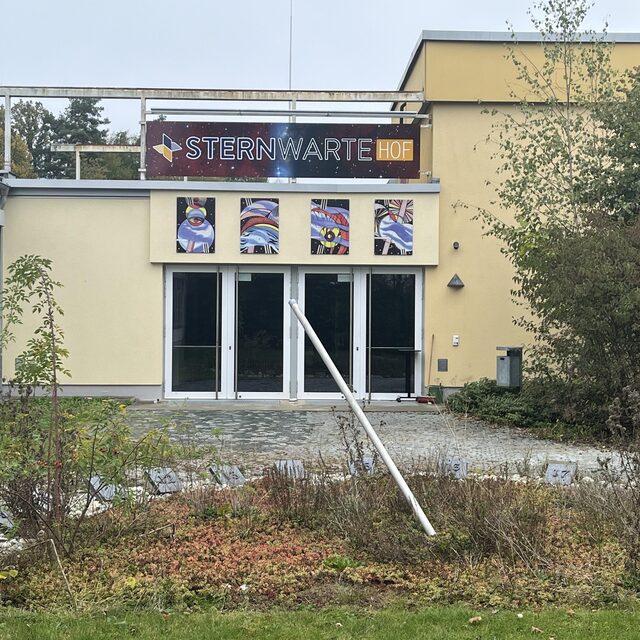 Eingangsbereich der Sternwarte Hof mit Sternwarte-Hof-Schild, Glastüren und bepflanztem Vorplatz